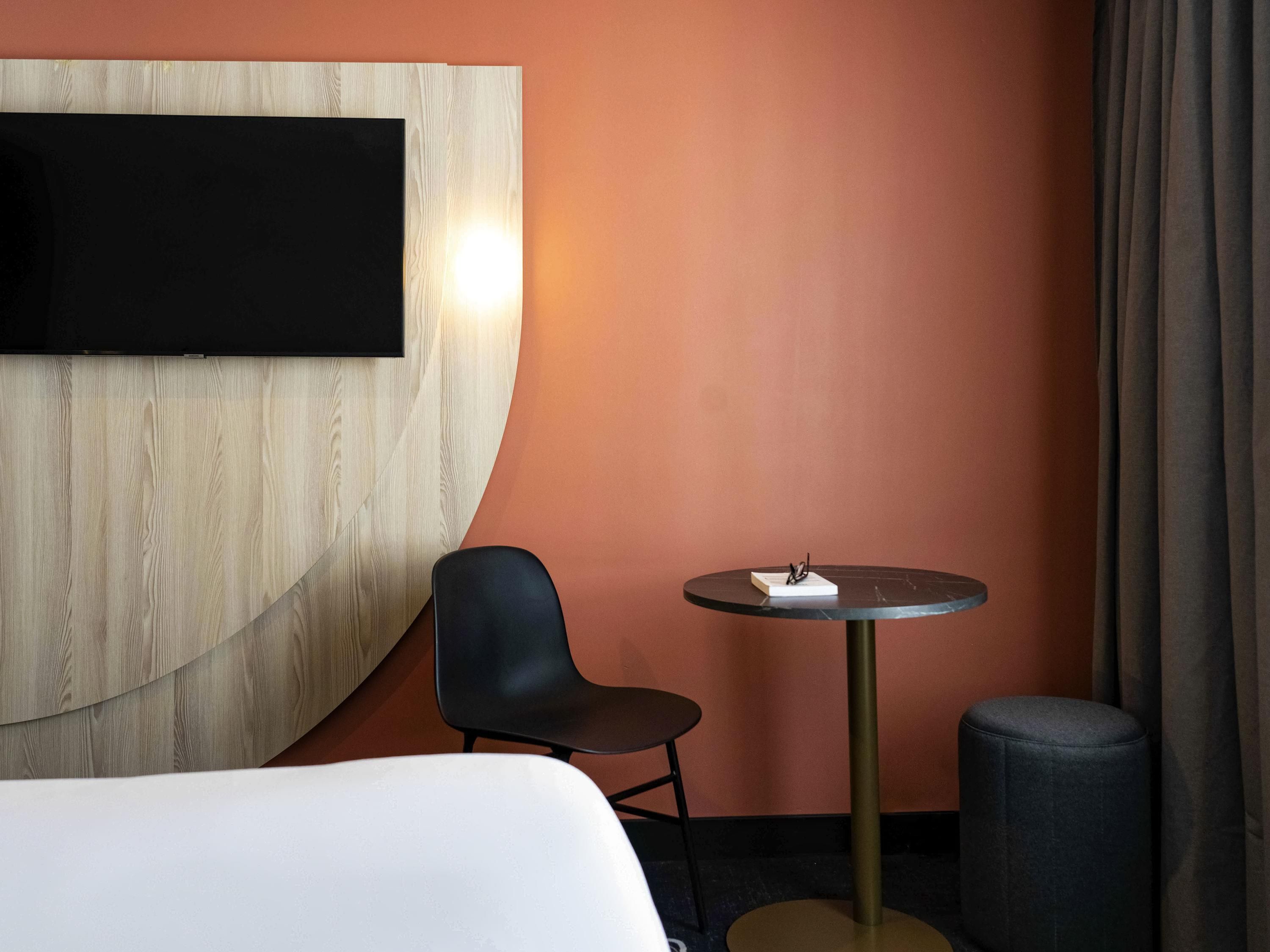Een hotelkamer met een groot bed, een klein rond tafeltje met een boek en glazen, een zwarte stoel en een tv bevestigd aan een houten paneel.