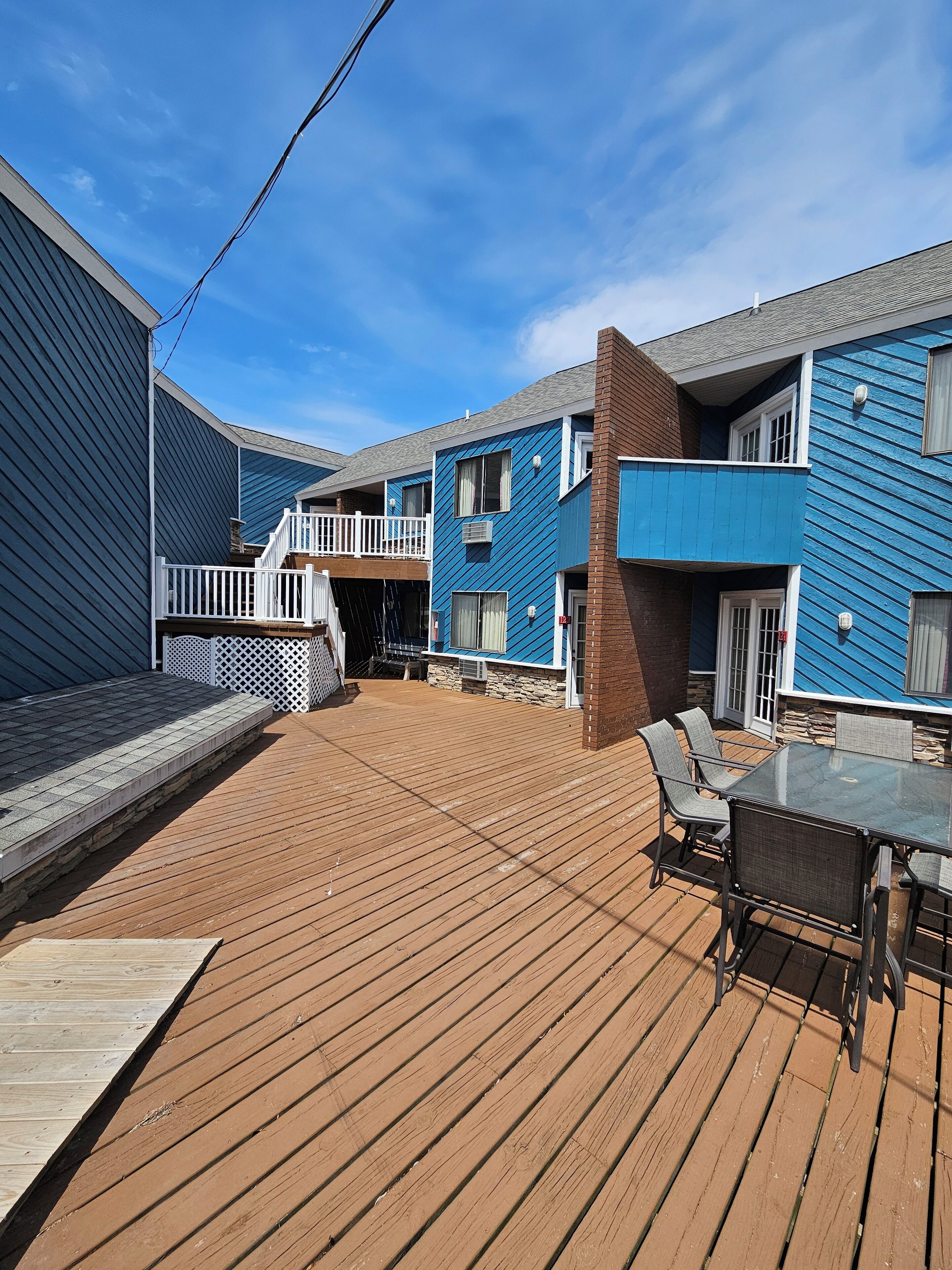 A wooden deck with a glass-top table and chairs, surrounded by blue buildings with balconies.