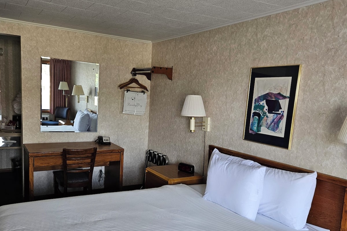 Une chambre d’hôtel avec un grand lit, un bureau avec une chaise, un miroir, une lampe fixée au mur et un tableau encadré.