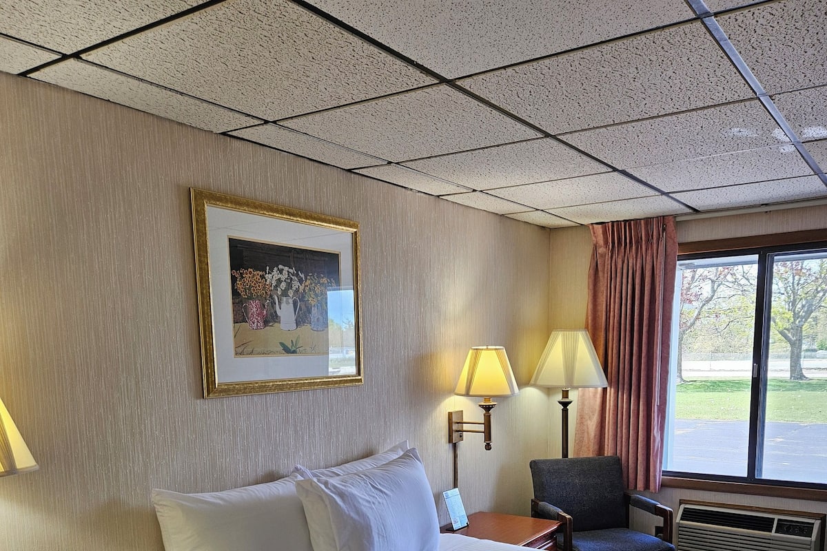Une chambre d’hôtel avec un grand lit, deux tables de chevet, une chaise, un bureau, une lampe, un tableau et une fenêtre avec des rideaux.