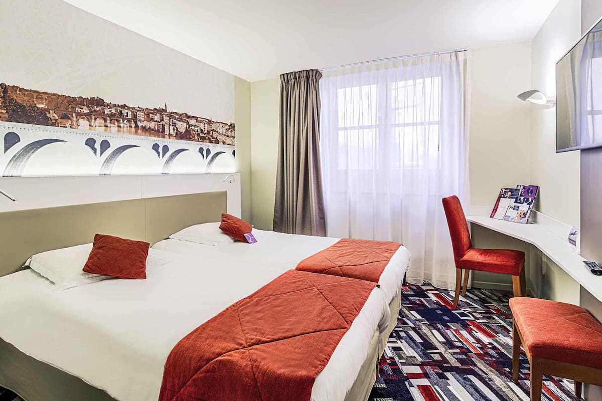 A hotel room with a bed, a desk, a chair, a TV, and a cityscape view.