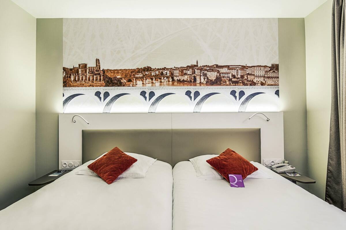 A hotel room with a cityscape view, two beds, a nightstand, and a window with a curtain.
