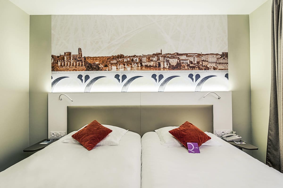 A hotel room with a cityscape view, two beds, a nightstand, and a window with a curtain.
