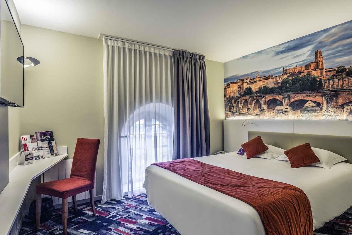 A hotel room with a bed, a desk, a chair, and a large window with curtains.
