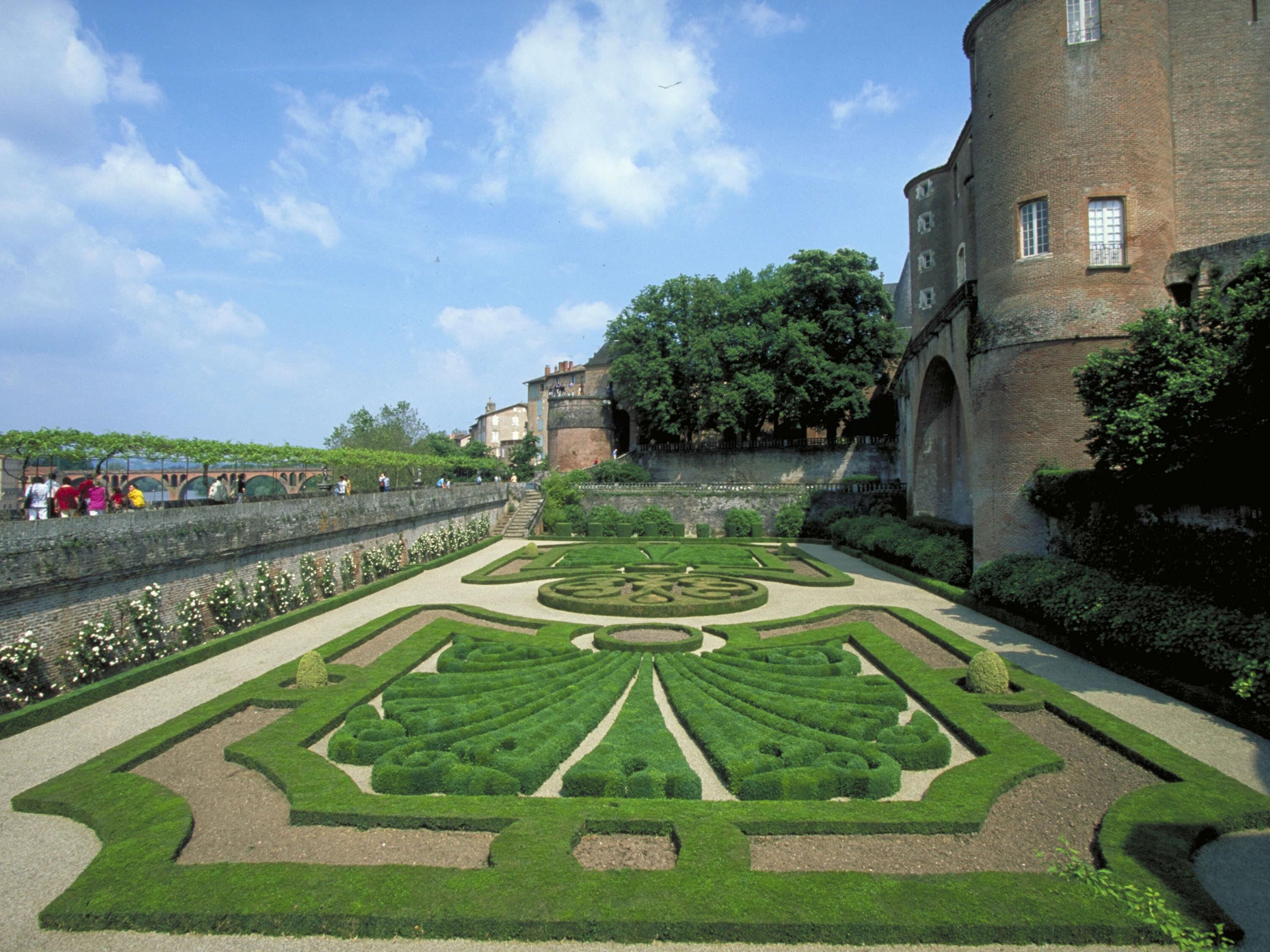 A formal garden with geometric patterns, a stone wall, and a historic building with arched windows.