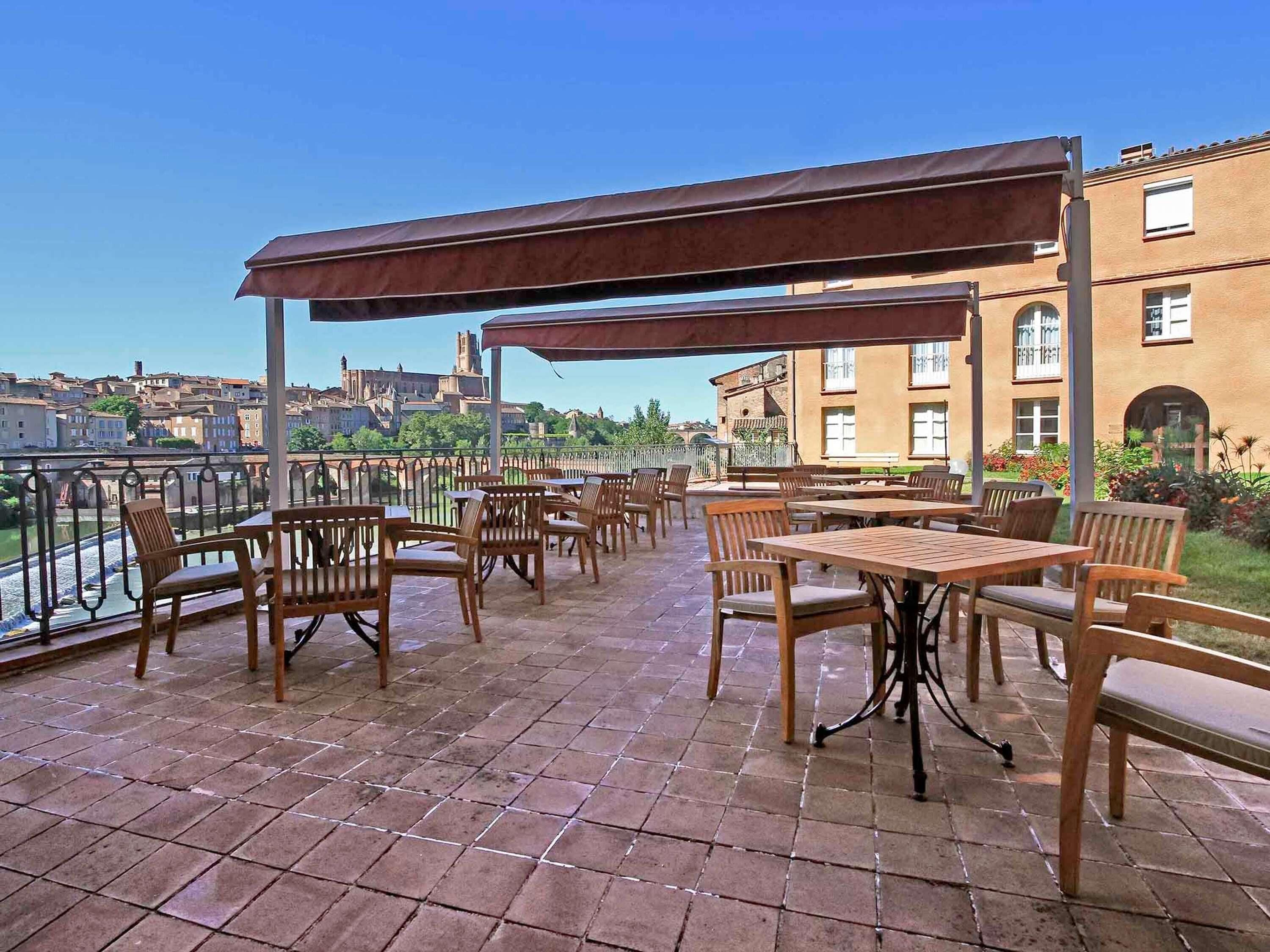 Een terras buiten met tafels en stoelen, een overdekt gedeelte en uitzicht op gebouwen en een rivier.