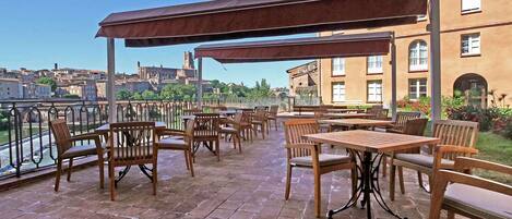 Una terraza al aire libre con mesas y sillas, una zona cubierta y vistas a edificios y un río.