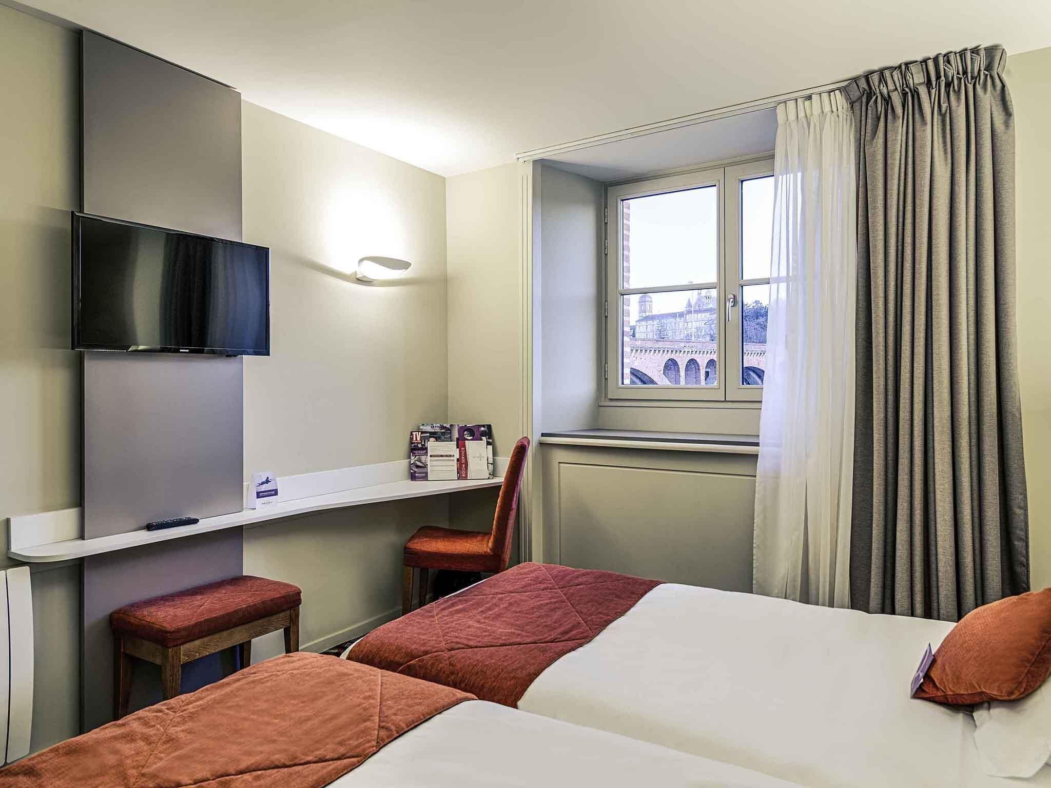 A hotel room with two beds, a TV mounted on the wall, a small desk with a chair, and a window with curtains.