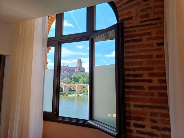A room with a large window, brick walls, and a view of a historic building and a bridge. - Mercure Albi Rives du Tarn (Albi)