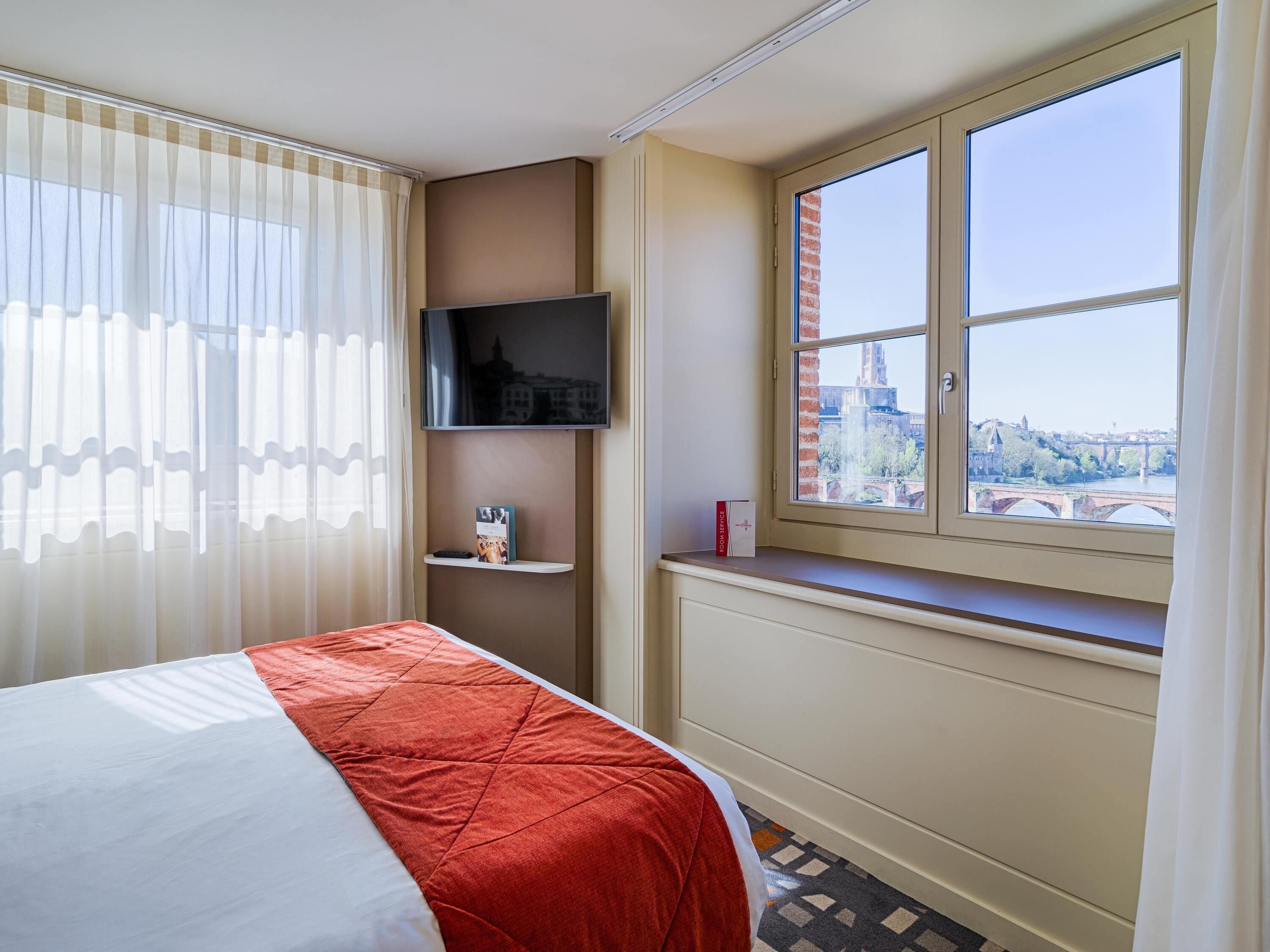 A hotel room with a bed, a TV mounted on the wall, a window with a view of a bridge and buildings, and a shelf with a book and a bottle.