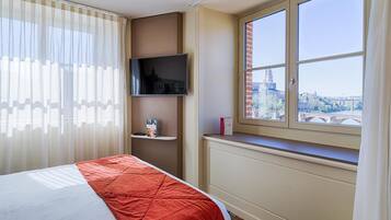 A hotel room with a bed, a TV mounted on the wall, a window with a view of a bridge and buildings, and a shelf with a book and a bottle.