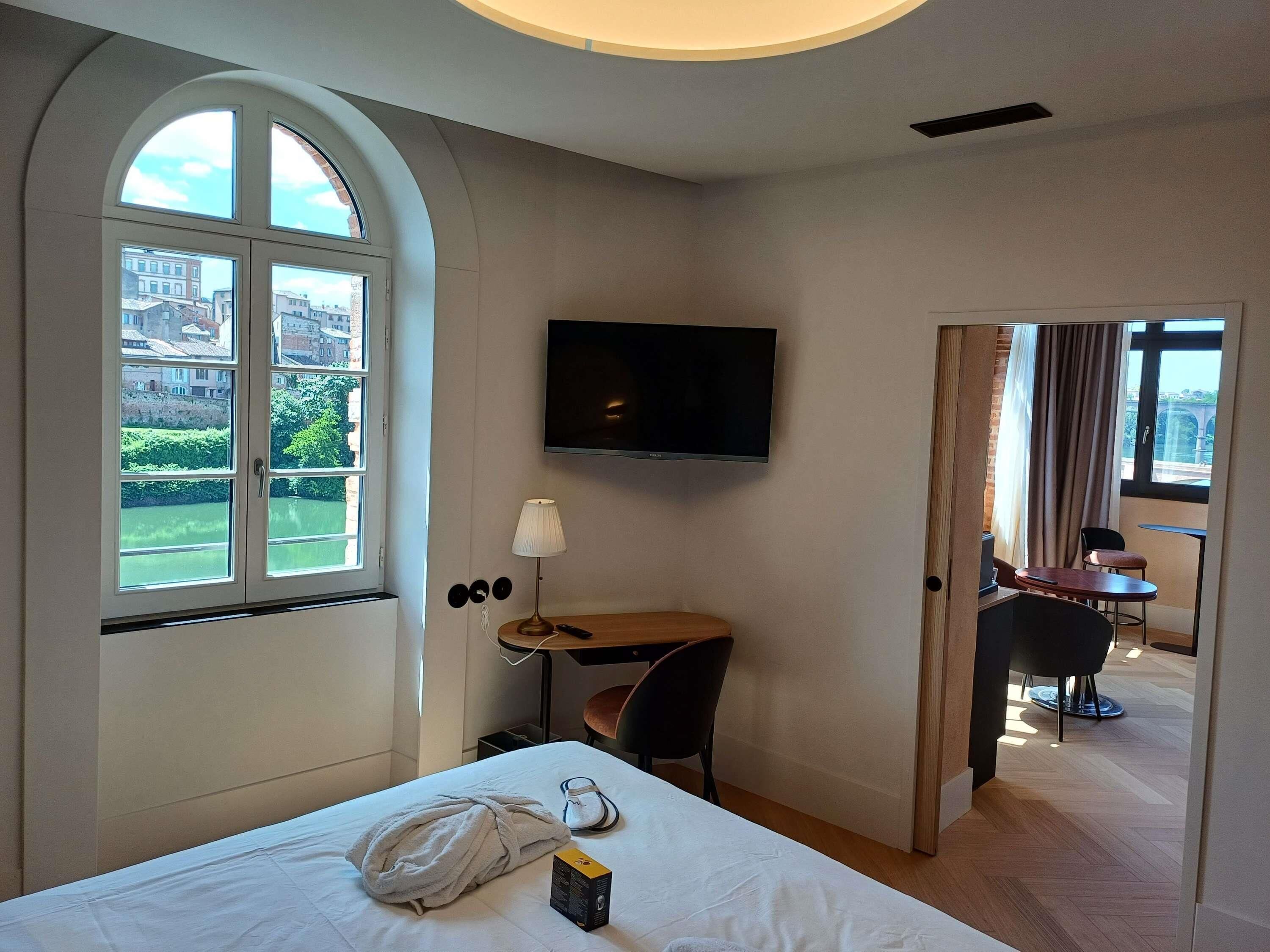 Une chambre d’hôtel équipée d’un lit, d’un bureau, d’une télévision et offrant une vue sur la ville grâce à de grandes fenêtres.