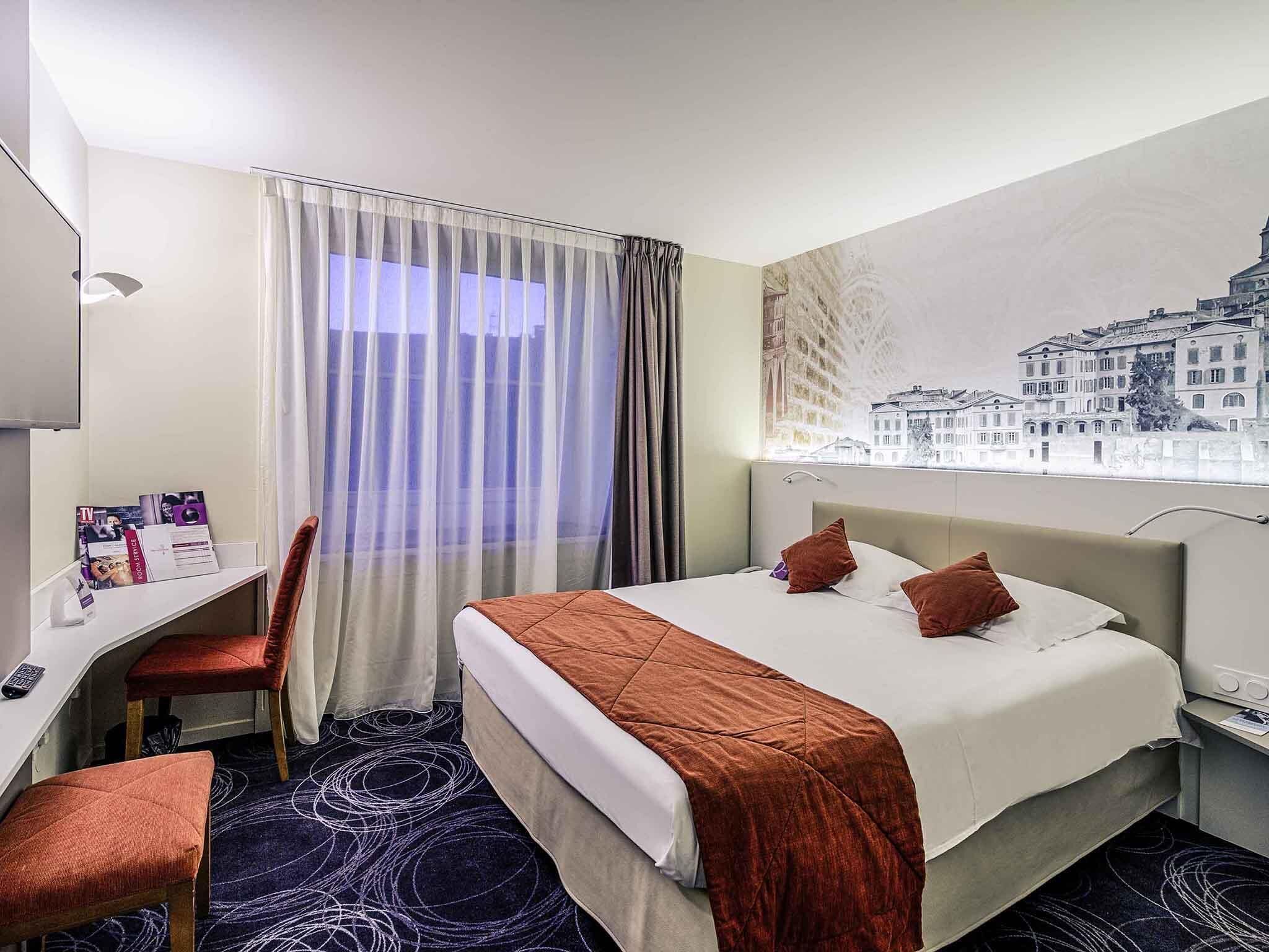 A hotel room with a bed, a desk, a chair, a window with a cityscape view, and a wall-mounted TV.