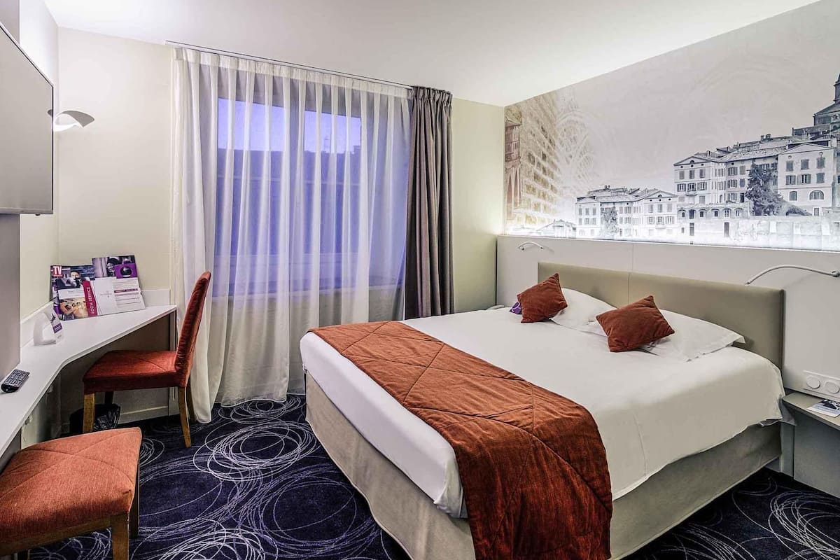 A hotel room with a bed, a desk, a chair, a window with a cityscape view, and a wall-mounted TV.