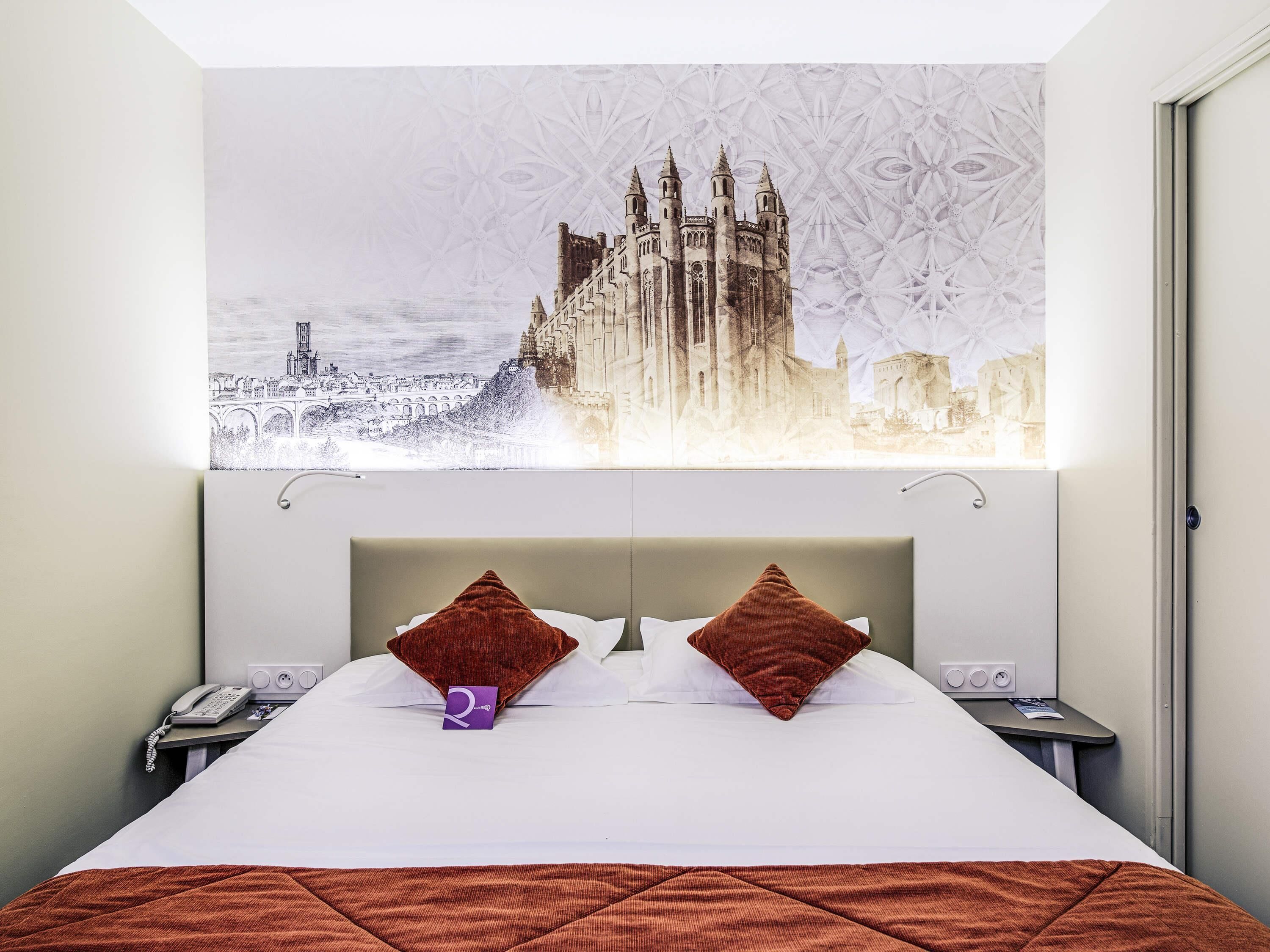 Een hotelkamer met een groot bed, twee kussens, een telefoon op het nachtkastje en een wandlamp.