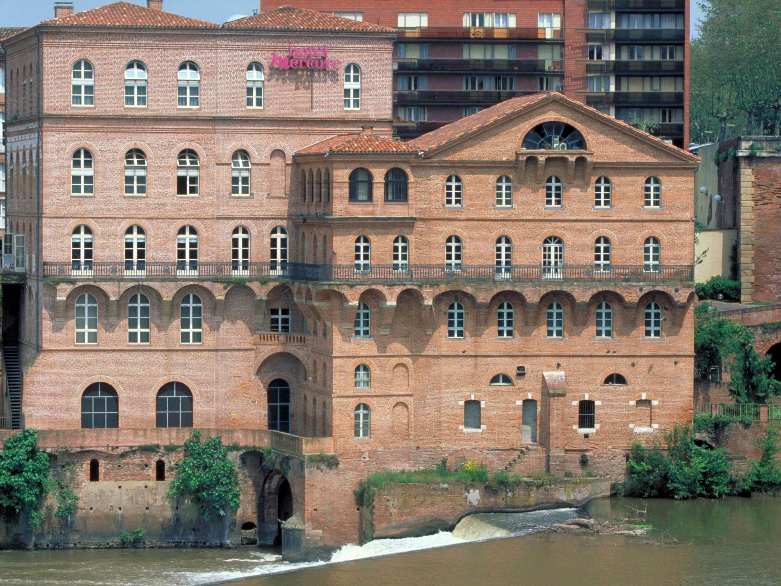 A historic brick building with arched windows and a central entrance, located near a river.