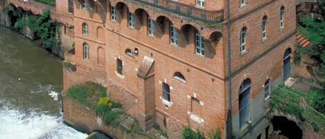 A multi-story brick building with arched windows and a balcony, adjacent to a river.