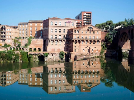 A historic building with arched windows and a reflection in the water.. Mercure Albi Rives du Tarn
