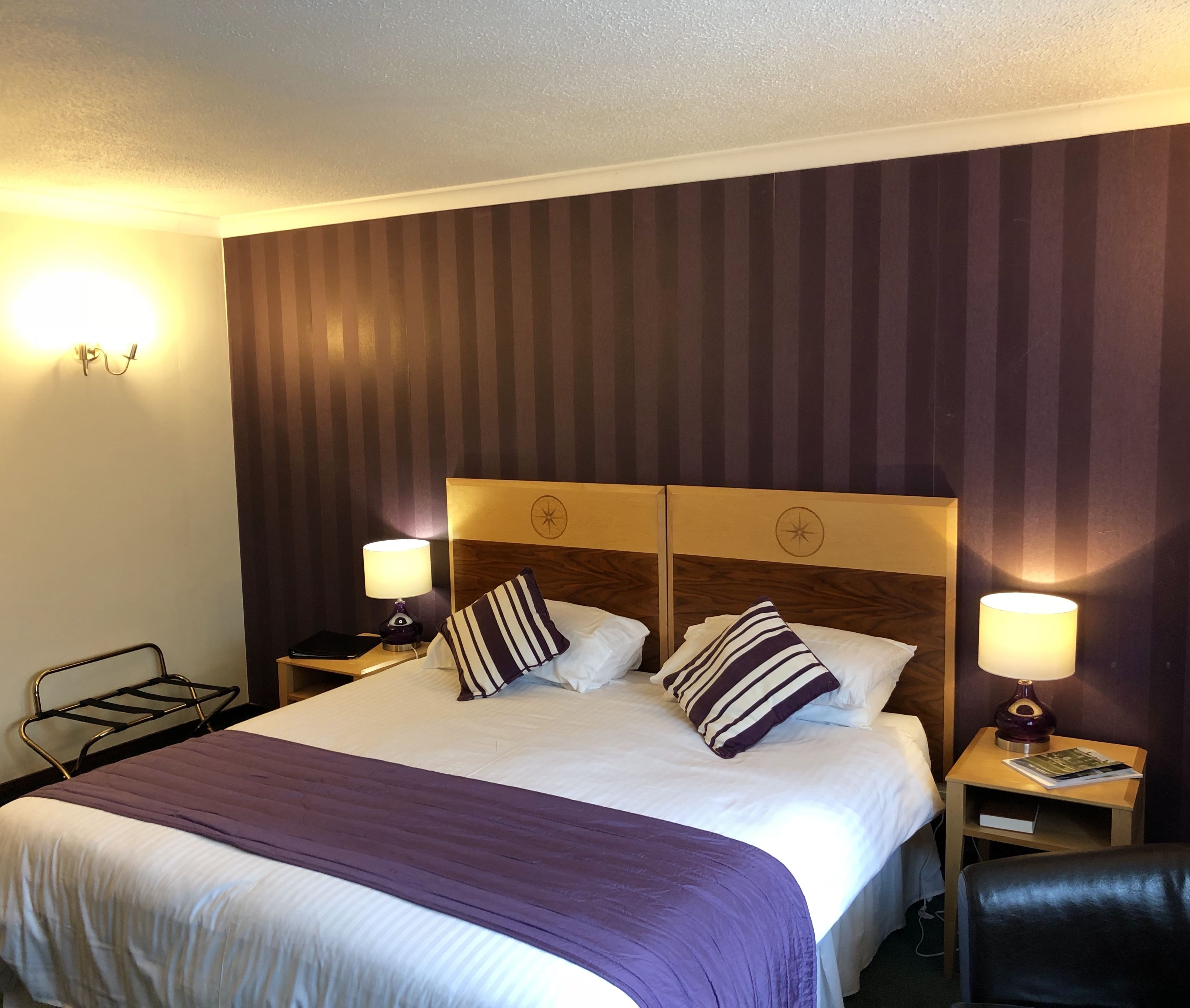 A hotel room with a large bed, two bedside tables with lamps, a headboard with a compass design, and a striped curtain.