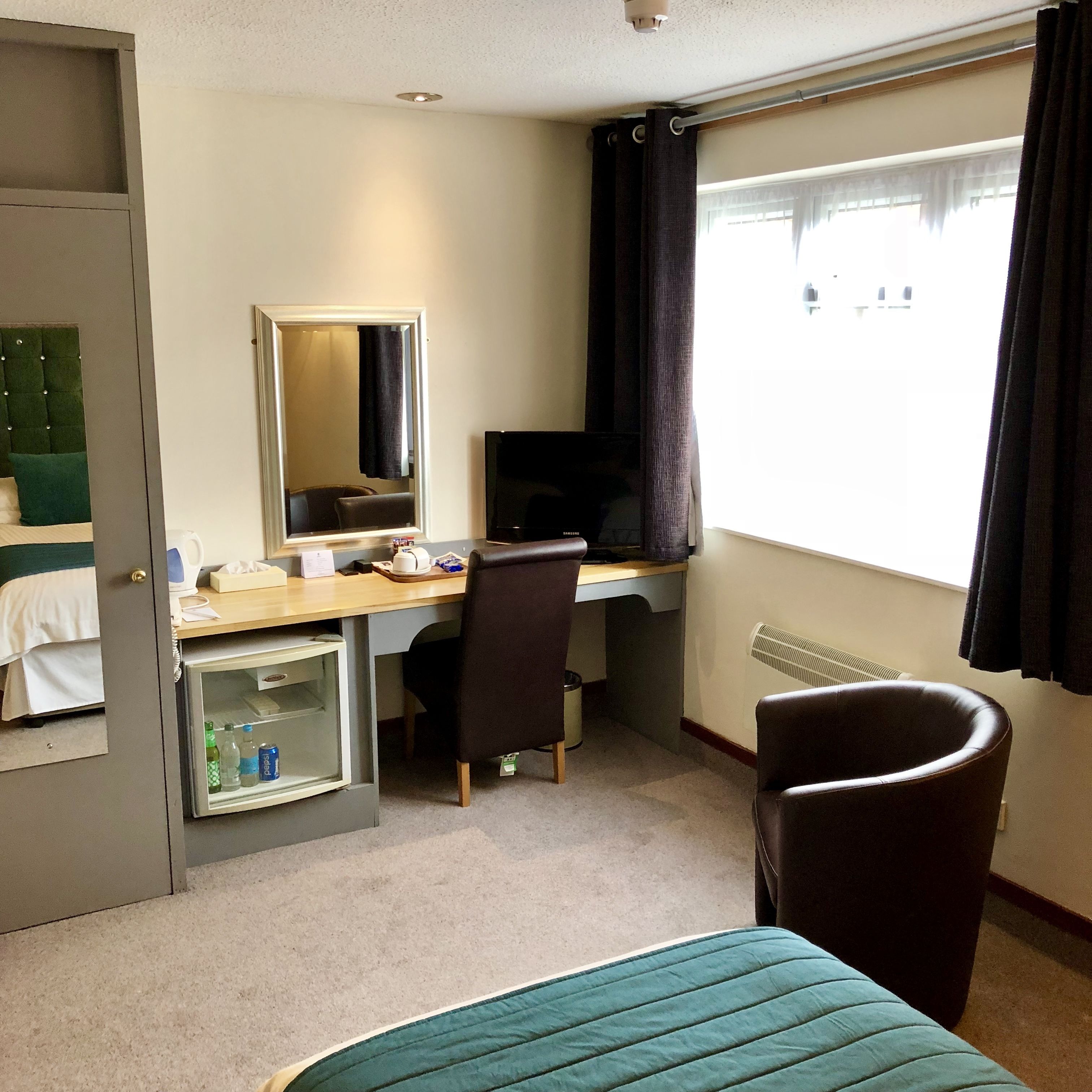 A hotel room with a bed, a desk with a computer, a chair, a minibar, and a window with curtains.