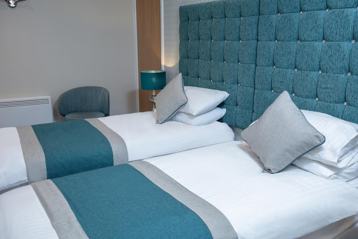 A hotel room with two beds, a teal upholstered headboard, a blue patterned chair, and a bedside table with a lamp.