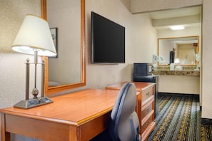 A hotel room with a wooden desk, a chair, a television, a mirror, and a bathroom area with a sink and mirror.