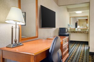 Habitación de hotel con un escritorio de madera, una silla, televisor, espejo y zona de baño con lavamanos y espejo.