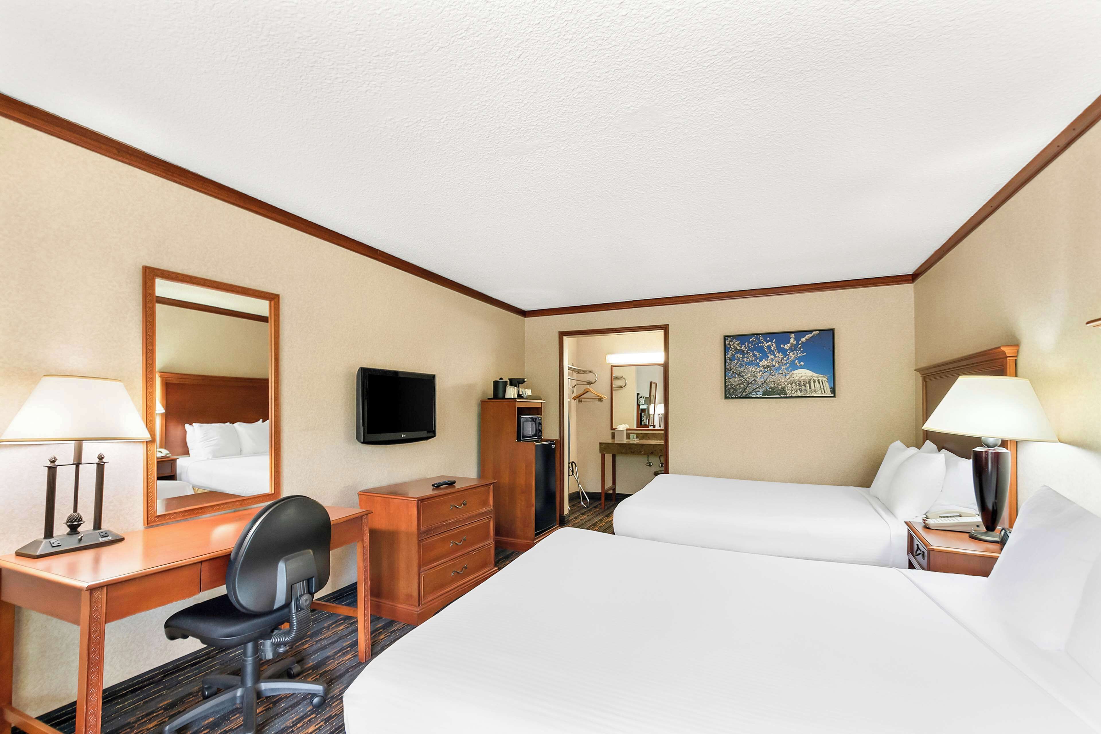 Habitación de hotel con dos camas, un escritorio con silla, cómoda, televisor y un cuadro en la pared.