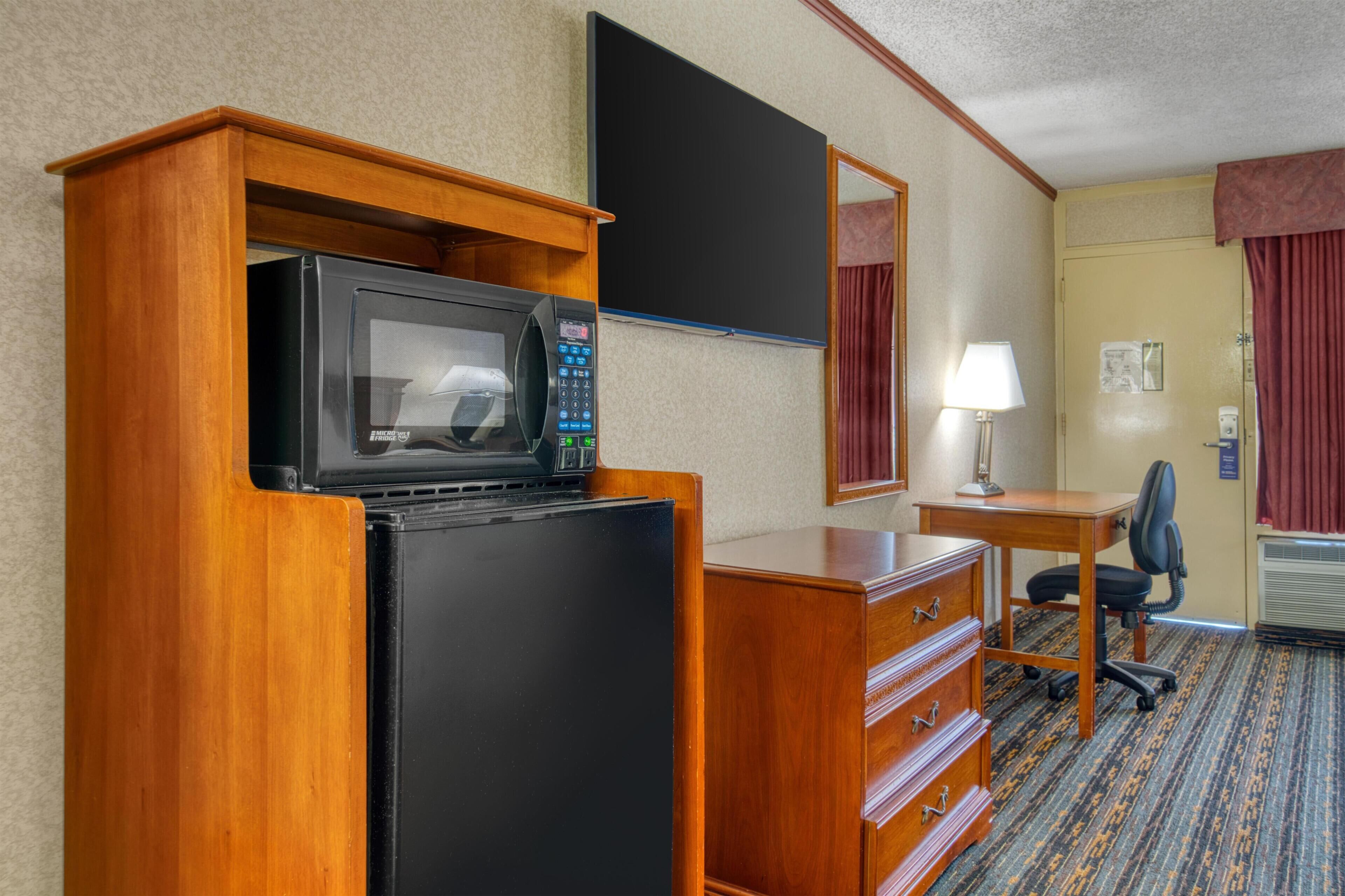 A hotel room with a wooden desk, a black refrigerator, a microwave, a chair, a lamp, and a mirror.