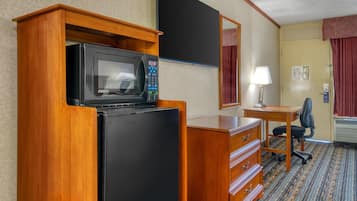 A hotel room with a wooden desk, a black refrigerator, a microwave, a chair, a lamp, and a mirror.