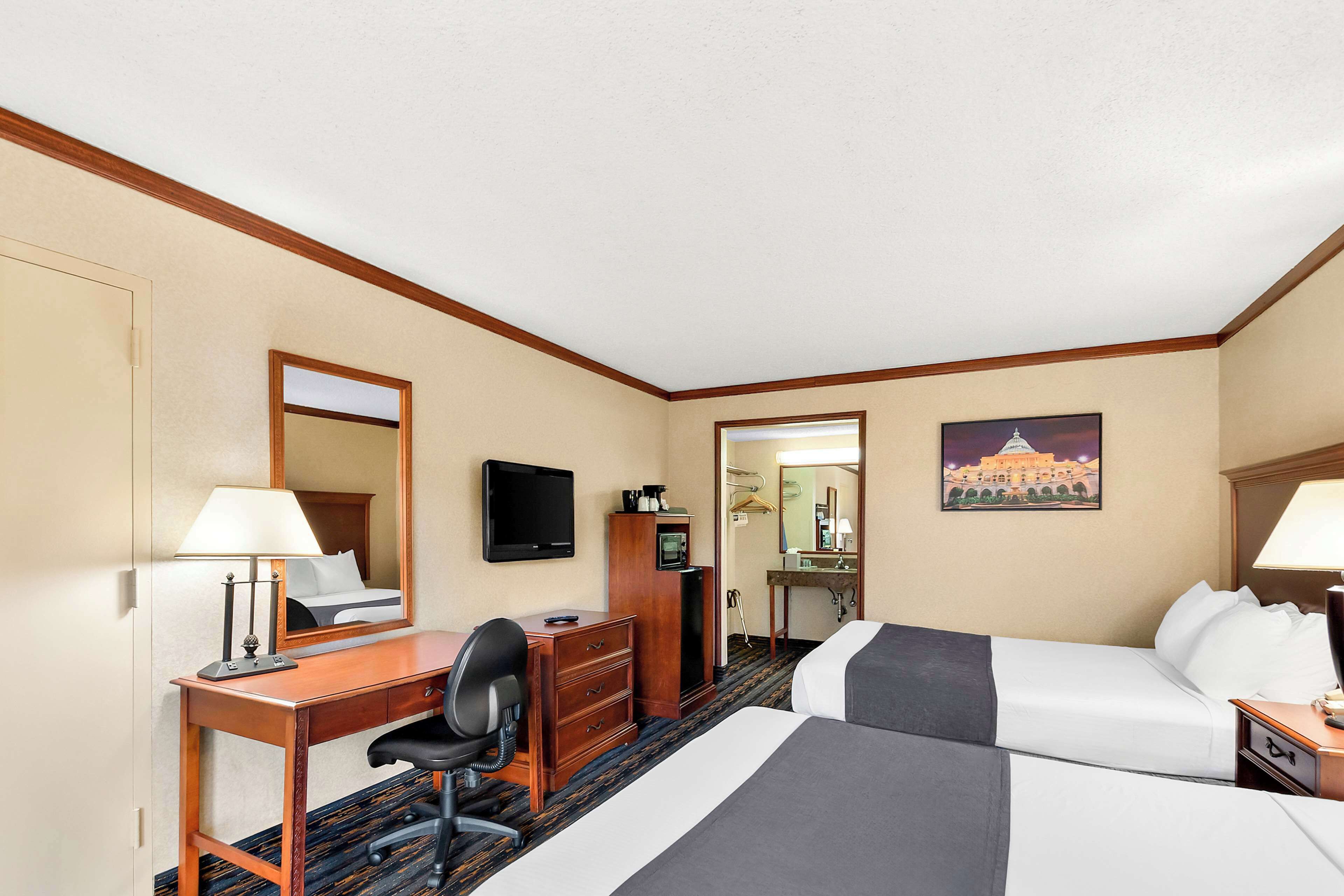A hotel room with two beds, a desk, a chair, a dresser, a TV, a painting, and a bathroom visible through an open door.