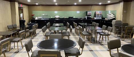 A cafeteria with multiple tables and chairs, a counter with food items, and a green wall with a sign.