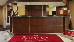 A reception desk at Ramada with a check-in counter, a red carpet with "RAMADA" and "Rochelle Park, NJ" text, and a "Manager on Duty" sign. - Ramada by Wyndham Rochelle Park Near Paramus (Rochelle Park)