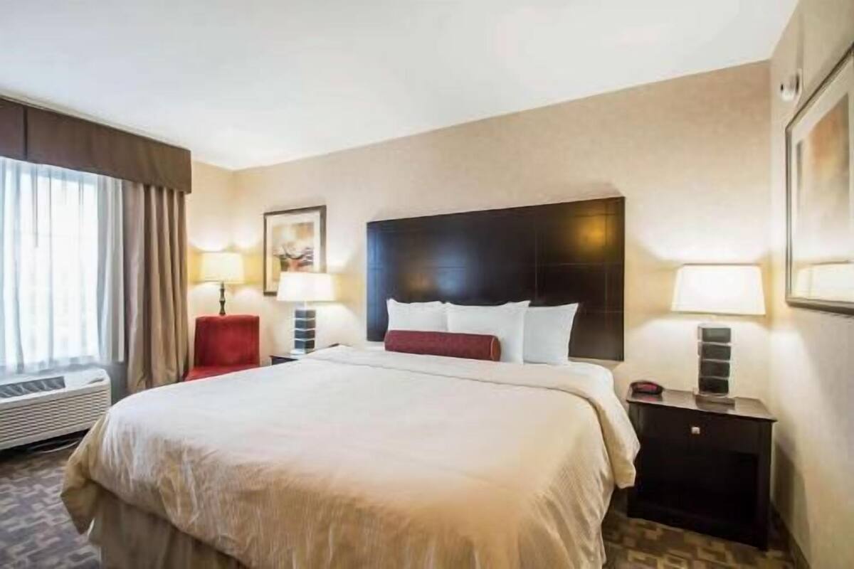 A hotel room with a large bed, two bedside tables, a red chair, a window with curtains, and a framed picture on the wall.