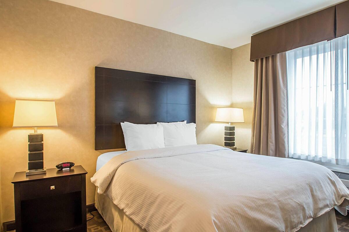 A hotel room with a large bed, two bedside tables with lamps, a digital clock, and a window with curtains.