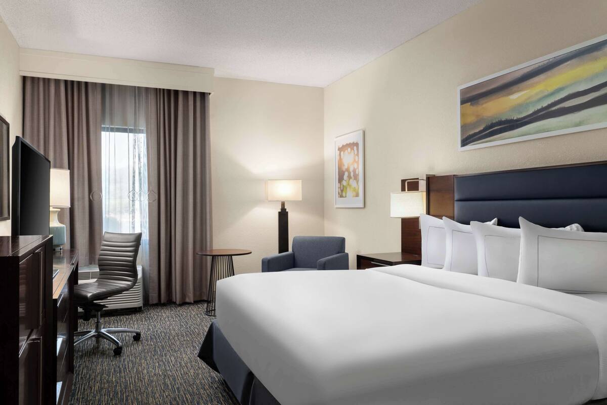 A hotel room with a large bed, a desk with a chair, a TV, a lamp, and a painting on the wall.