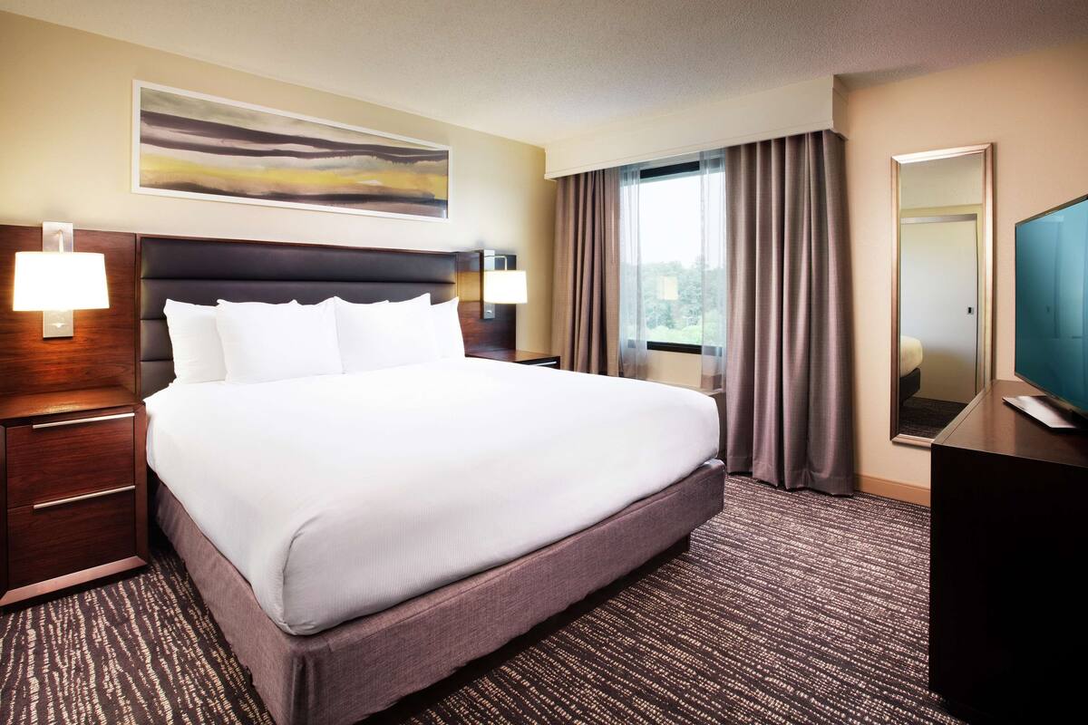 A hotel room with a large bed, a nightstand with a lamp, a desk with a computer, and a window with curtains.