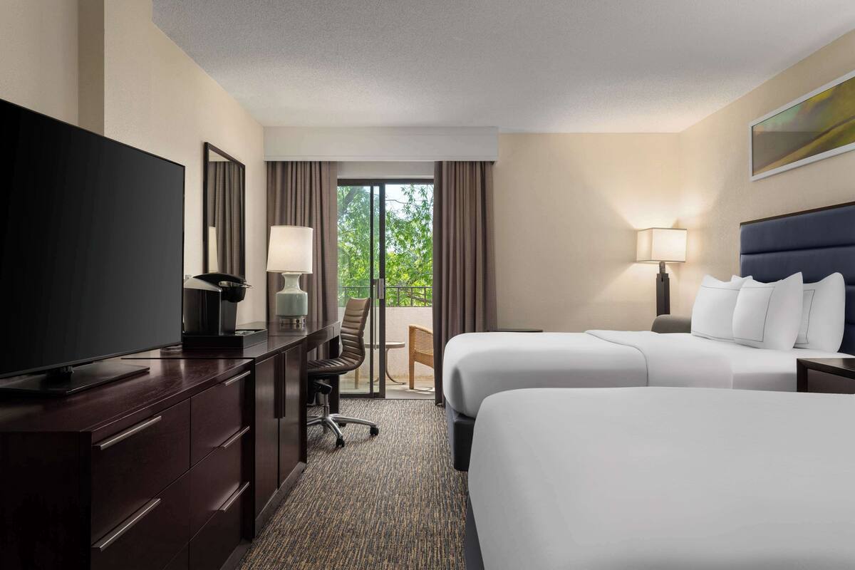 A hotel room with two beds, a desk with a TV, a chair, and a view of the outdoors.