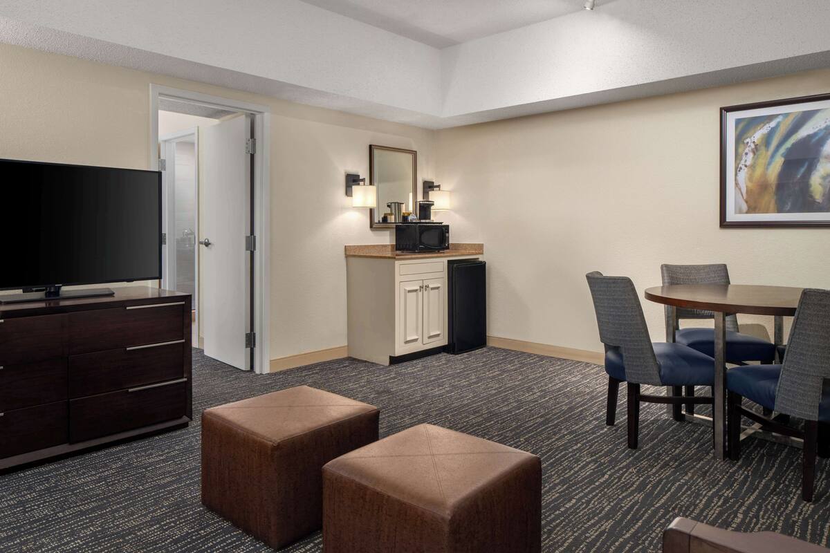 A hotel room with a television, a minibar, a dining area with chairs, and a carpeted floor.