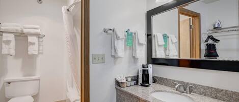 A bathroom with a toilet, sink, and shower area. There are towels hanging on a rack, a coffee maker, and a trash can.