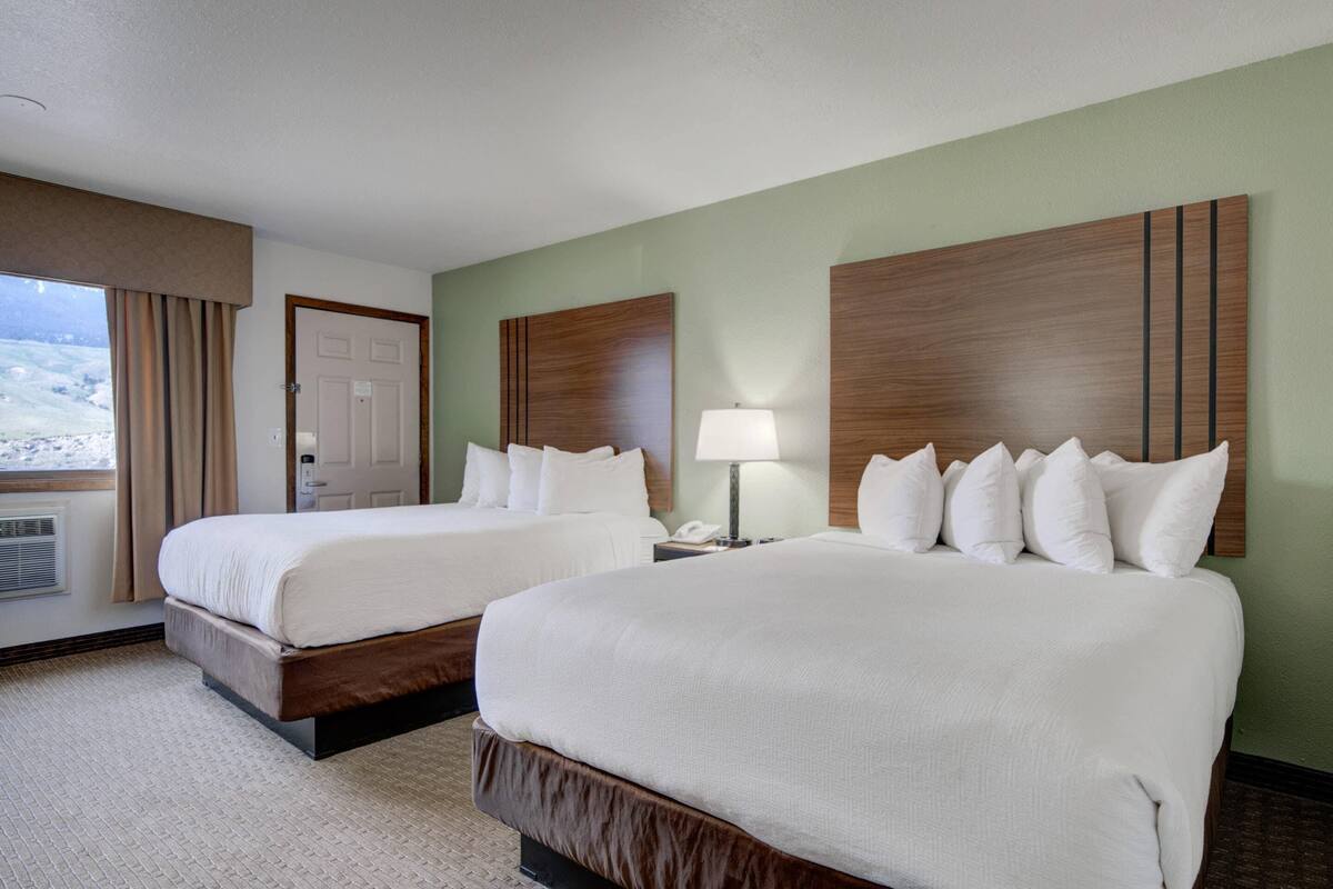 A hotel room with two beds, a window with curtains, a door, and a lamp on a nightstand.