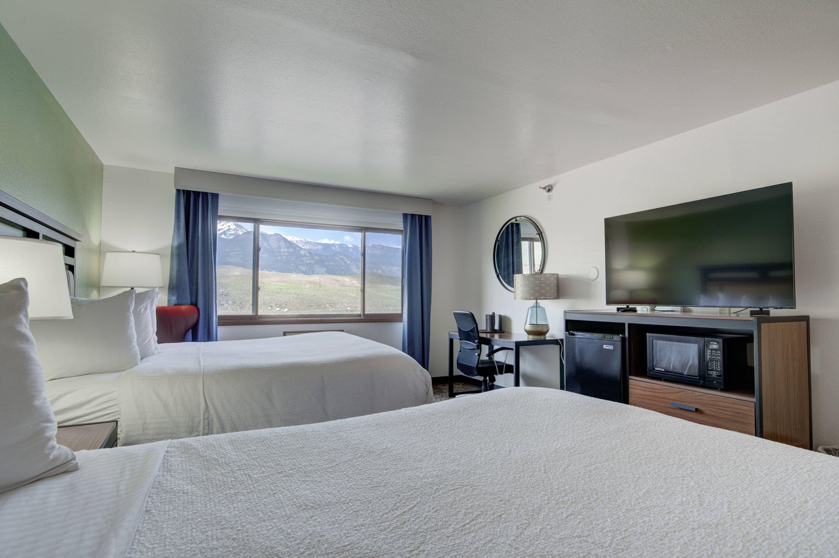 A hotel room with two beds, a desk with a chair, a television, a microwave, and a window with a mountain view.