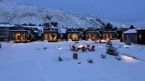 Une rangée de chalets enneigés, dotés de sièges extérieurs, avec une chaîne de montagnes en arrière-plan.
