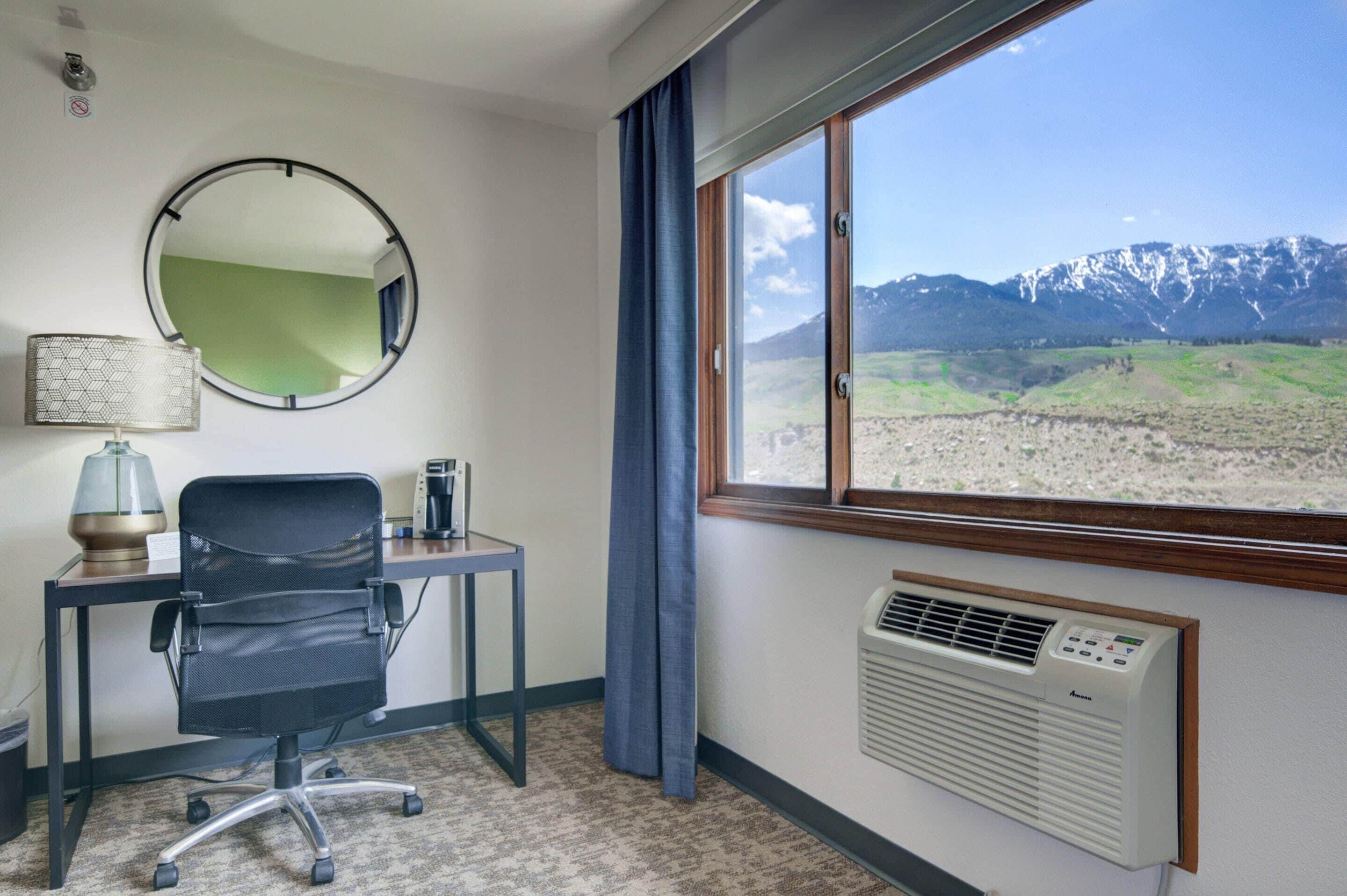 A hotel room with a desk, chair, mirror, lamp, air conditioning unit, and a window with a mountain view.