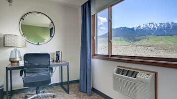 A hotel room with a desk, chair, mirror, lamp, air conditioning unit, and a window with a mountain view.