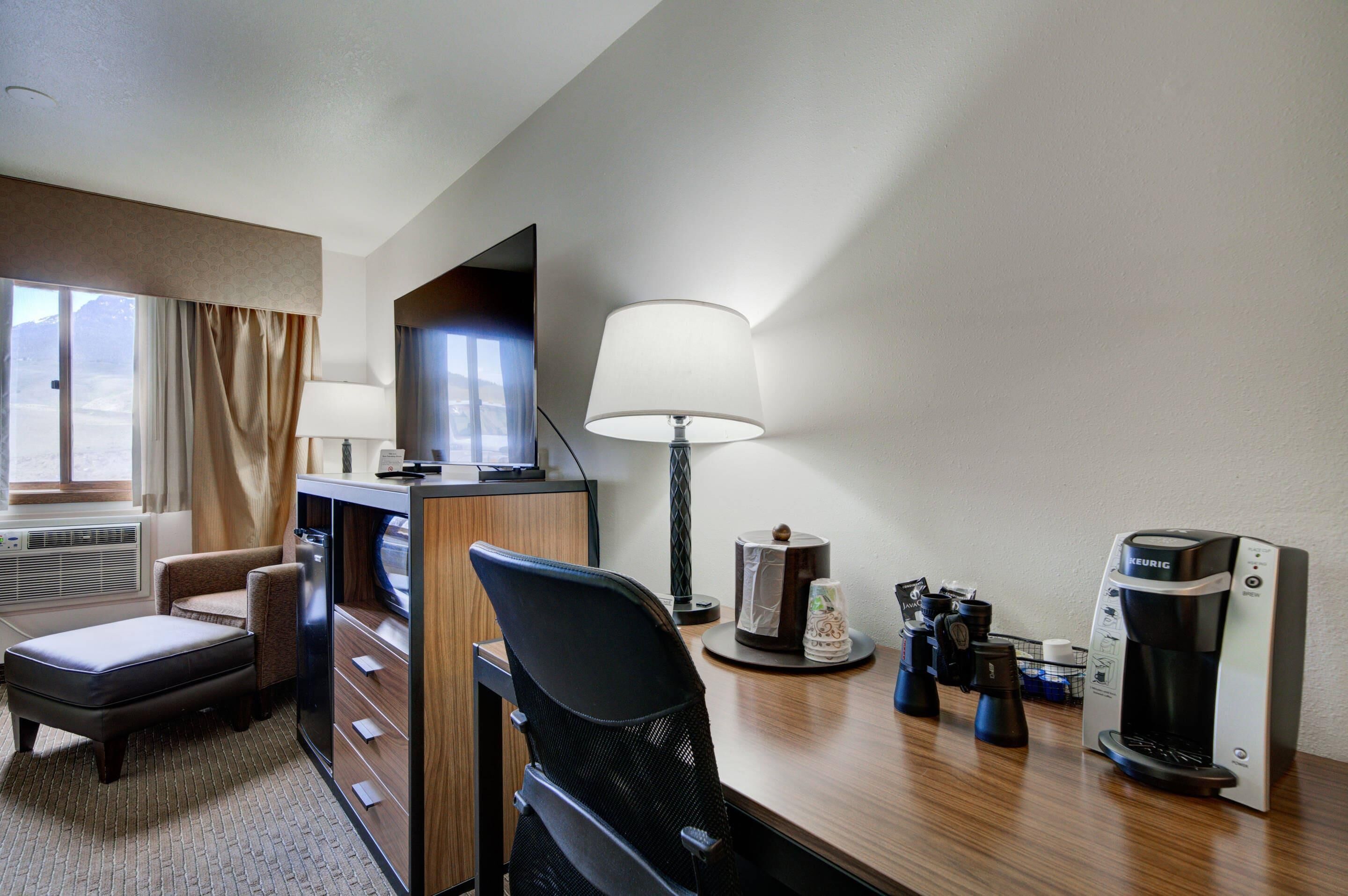 A hotel room with a desk, a chair, a television, a lamp, a coffee maker, and a window with curtains.