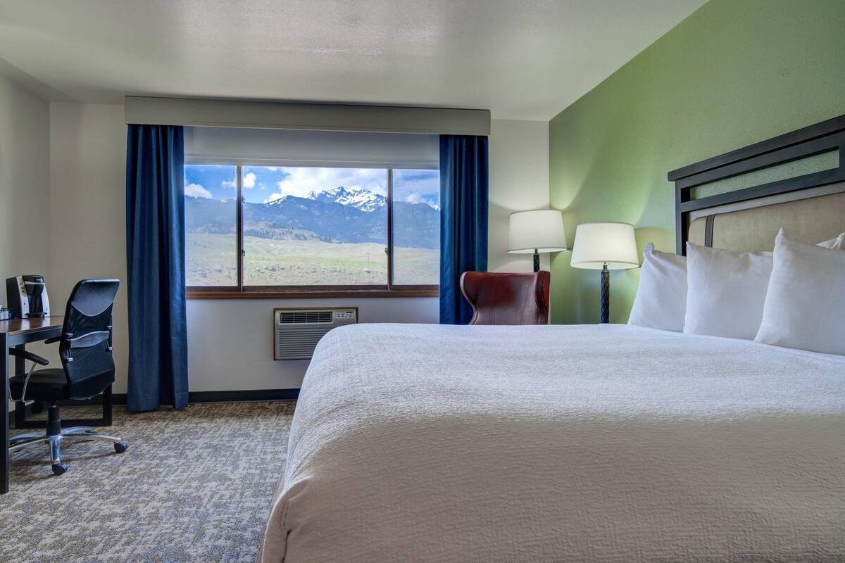 A hotel room with a large bed, a desk with a chair, a lamp, and a view of mountains through a window.