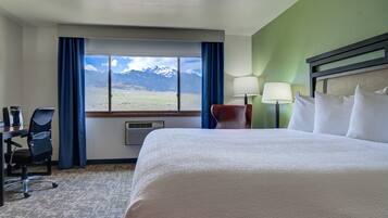 A hotel room with a large bed, a desk with a chair, a lamp, and a view of mountains through a window.