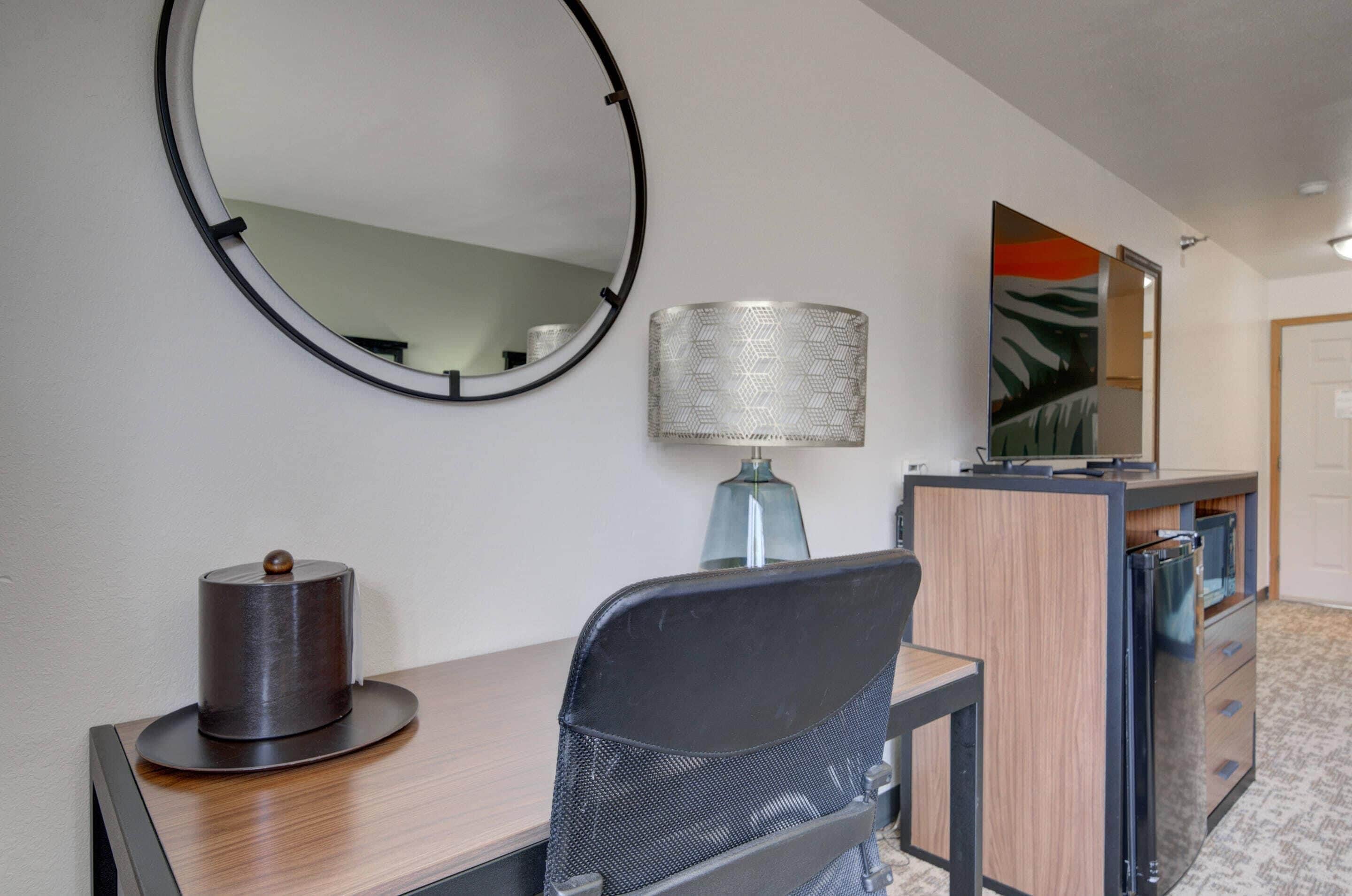 Habitación de hotel con un espejo redondo, una lámpara, un escritorio con computadora, una silla, un televisor y un minibar.