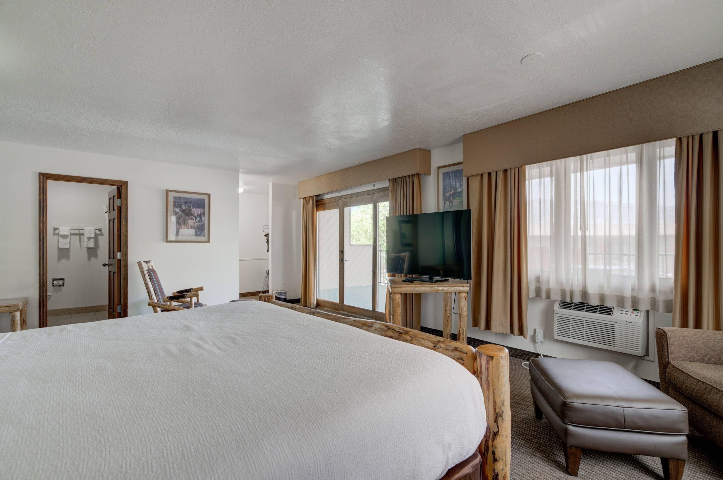 A hotel room with a large bed, a television on a wooden stand, a chair, and an air conditioning unit.